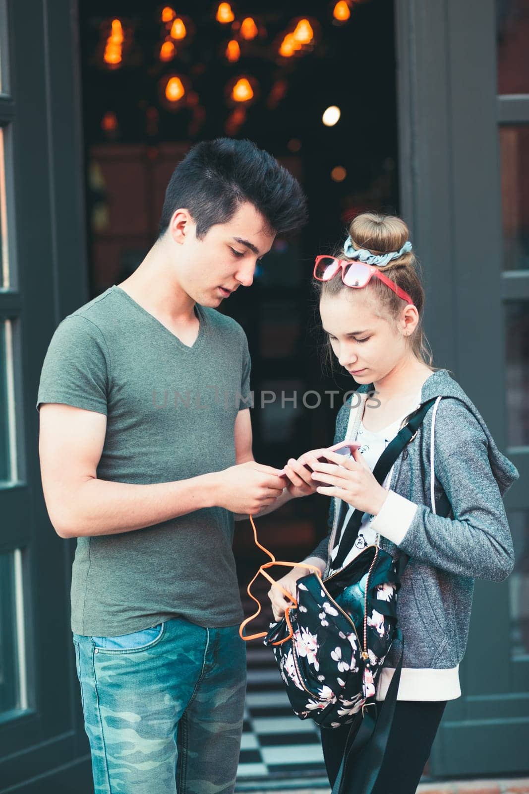 Couple of friends, teenage girl and boy,  using smartphones, talking together, standing on street in center of town, spending time together