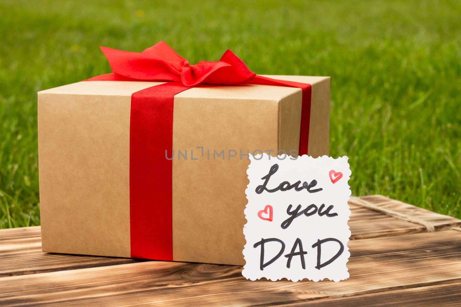 Greeting card Love You Dad with a gift box on a rustic wooden tabletop and green grass background