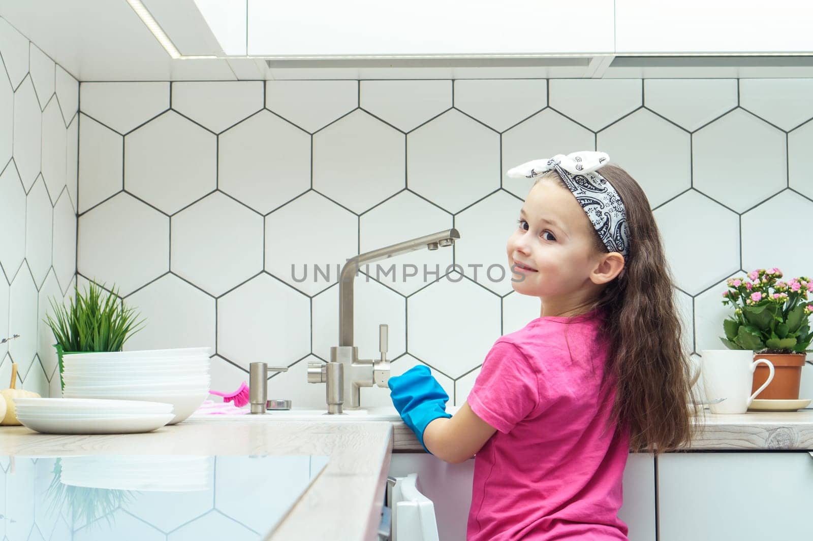 Back view of happy little preteen girl with long dark hair wearing pink T-shirt, big blue rubber gloves, standing near sink, preparing for washing dishes in kitchen. Housework, cleaning, dishwashing.