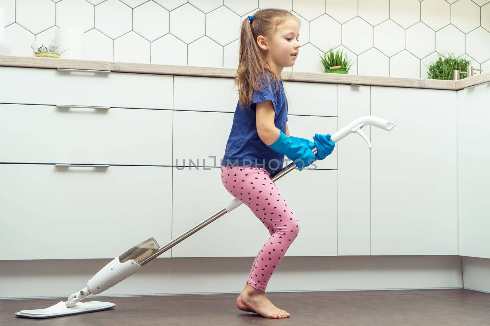 Portrait of funny little preteen girl with long dark hair wearing blue T-shirt, pink leggings, blue rubber gloves, riding on handheld spray mop as witch near table in kitchen. Housework, housekeeping.