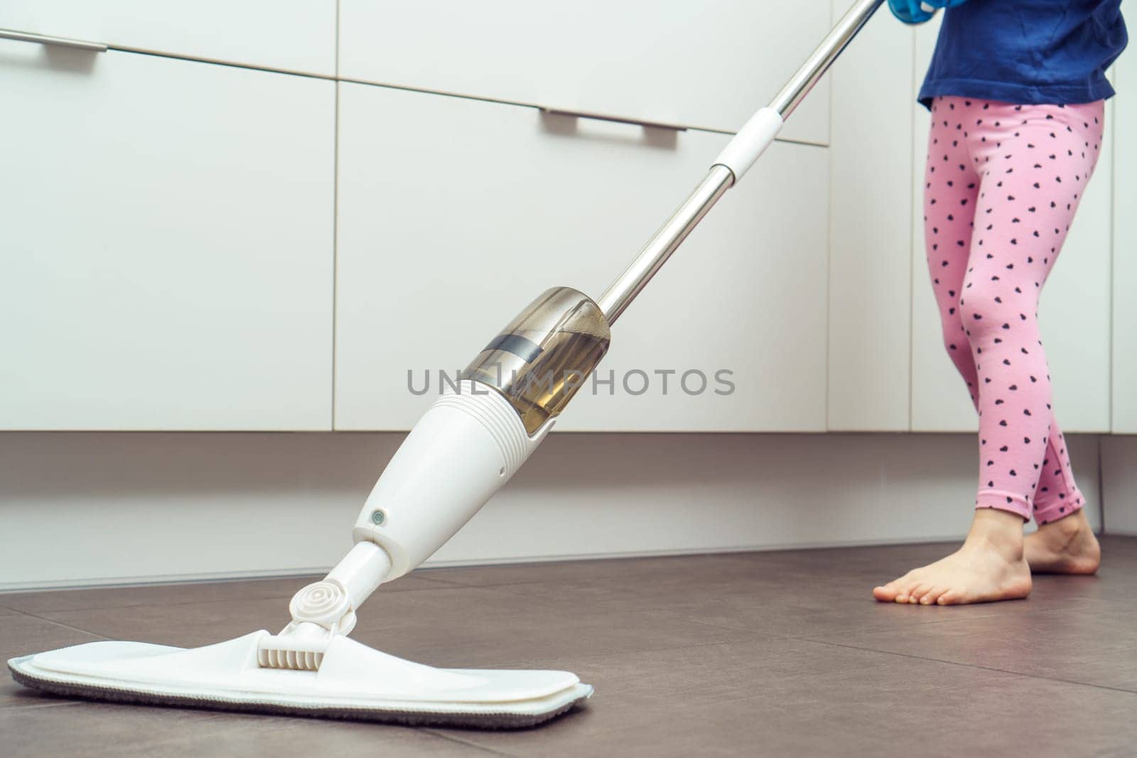 Cropped photo of little preteen girl wearing blue T-shirt, pink leggings, holding handheld spray mop, washing cleaning floor with microfiber cloth, spraying bottle in kitchen. Housework, housekeeping.