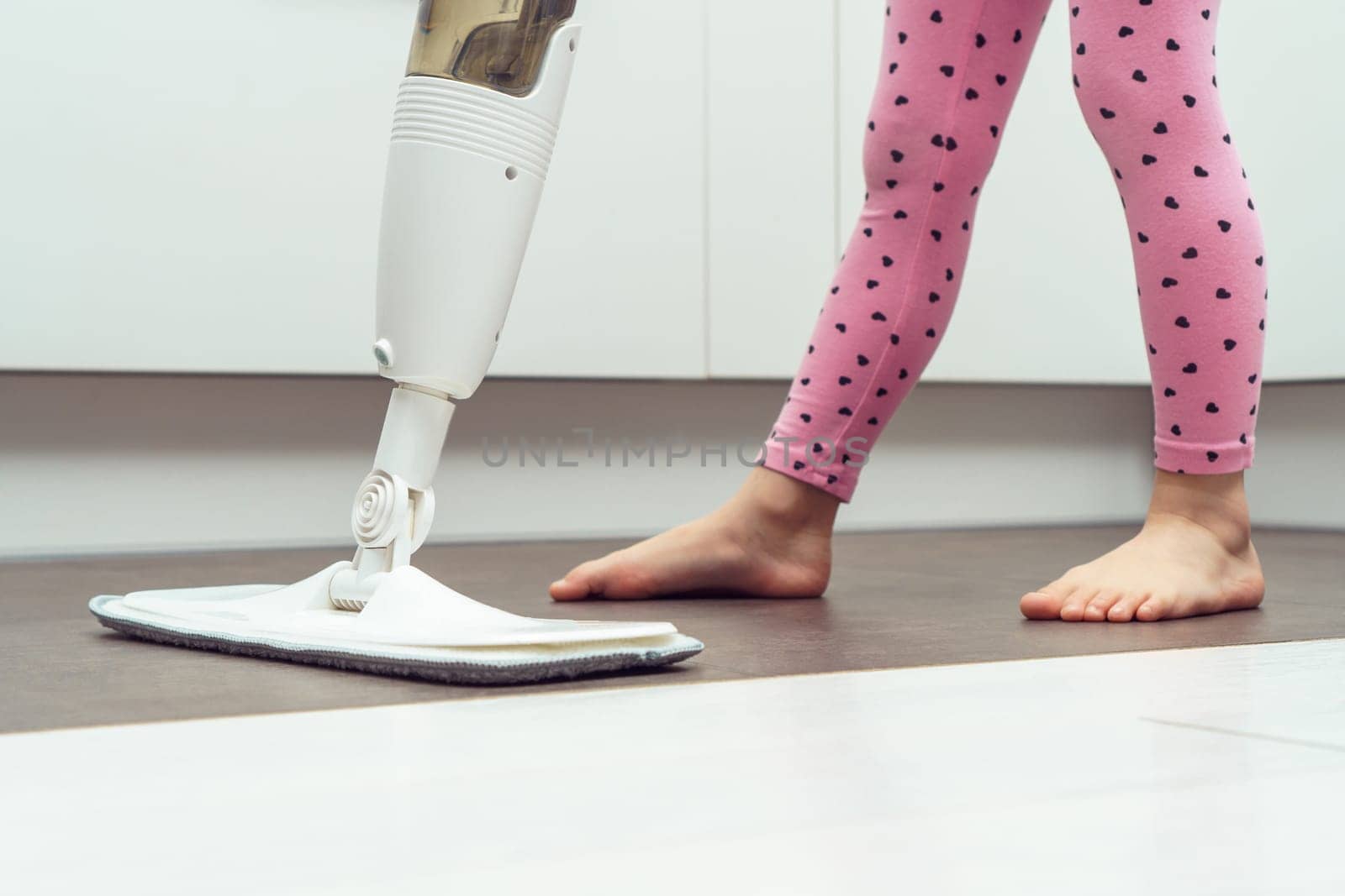 Unrecognizable little preteen girl wearing blue T-shirt, pink leggings, holding white handheld spray mop, mopping floor with microfiber cloth, refillable bottle in kitchen. Housework, housekeeping.
