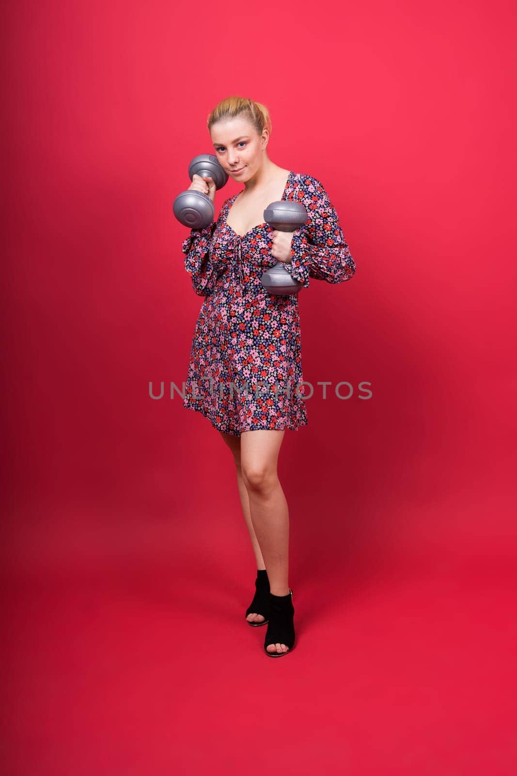 Beautiful model in sportswear with dumbbells looks at camera on yellow, red and dark background