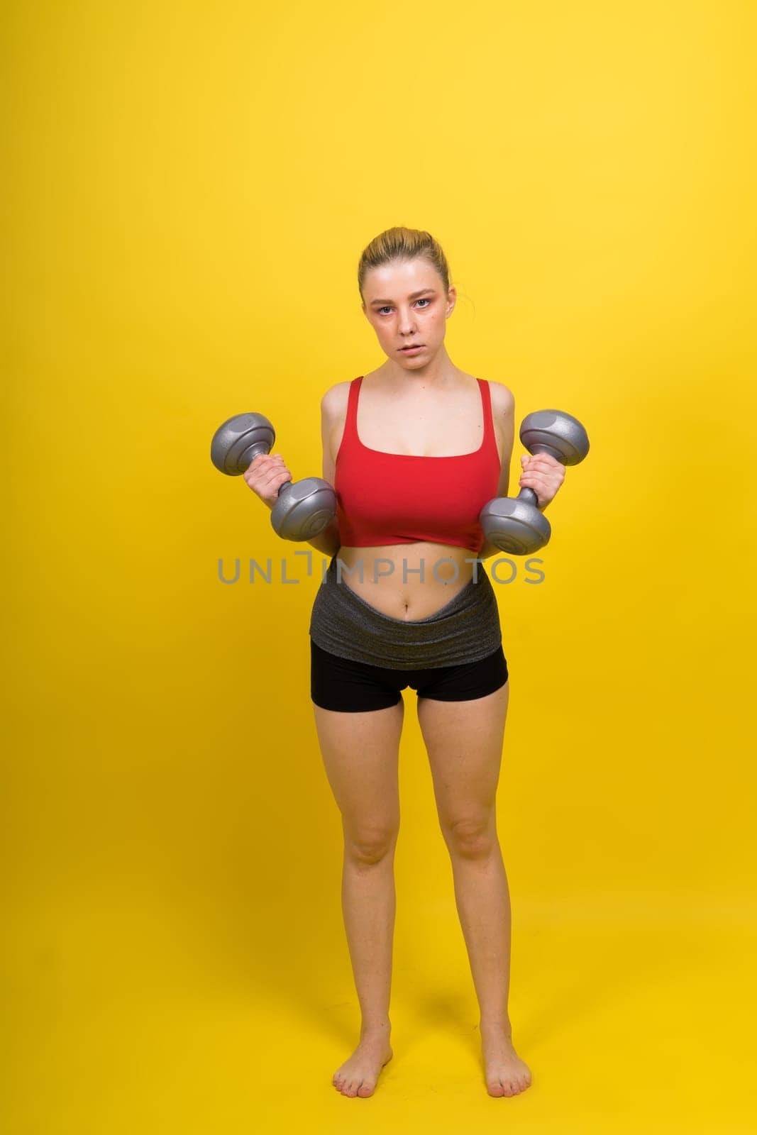 Beautiful model in sportswear with dumbbells looks at camera on yellow, red and dark background
