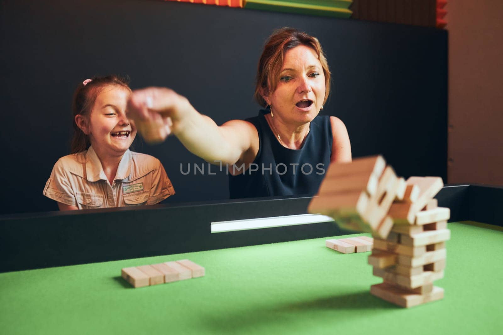 Excited woman playing jenga game with her daughter in play room. Woman removing block from stack. Falling tower of wooden blocks. Game of skill and fun. Family time