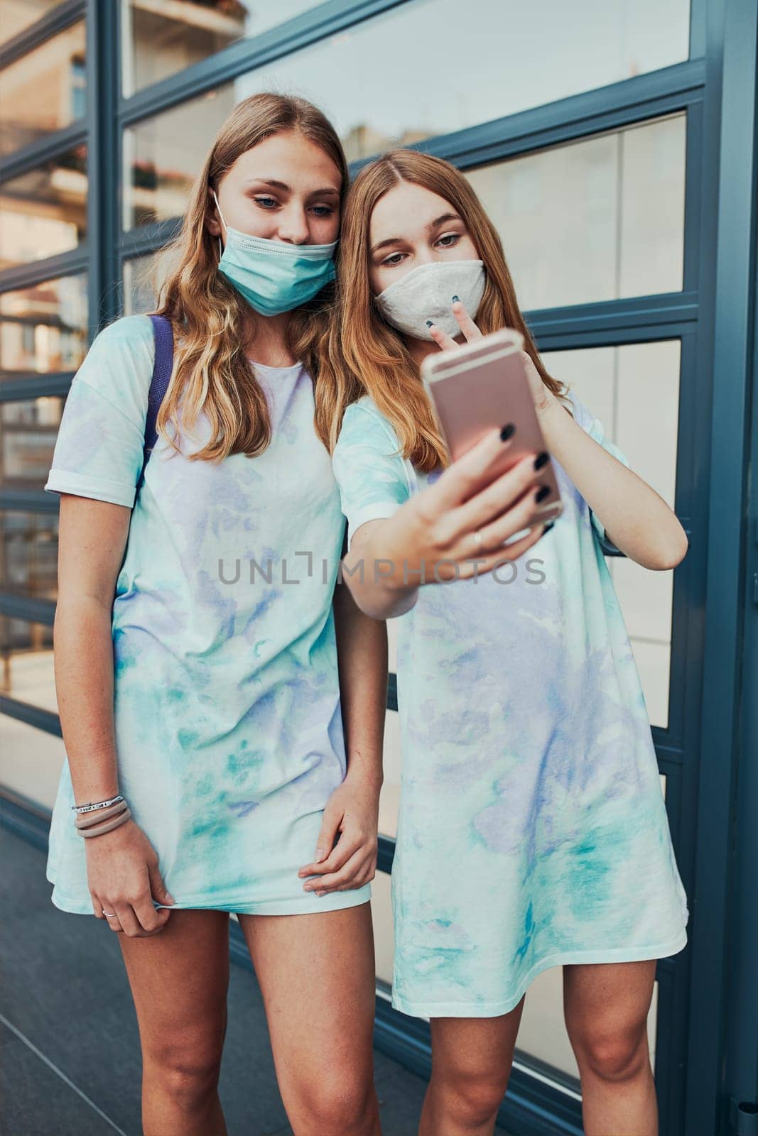 Young women girls taking selfie photos using smartphone standing at store front downtown. Girls wearing face masks to avoid virus infection and to prevent the spread of disease in time of coronavirus