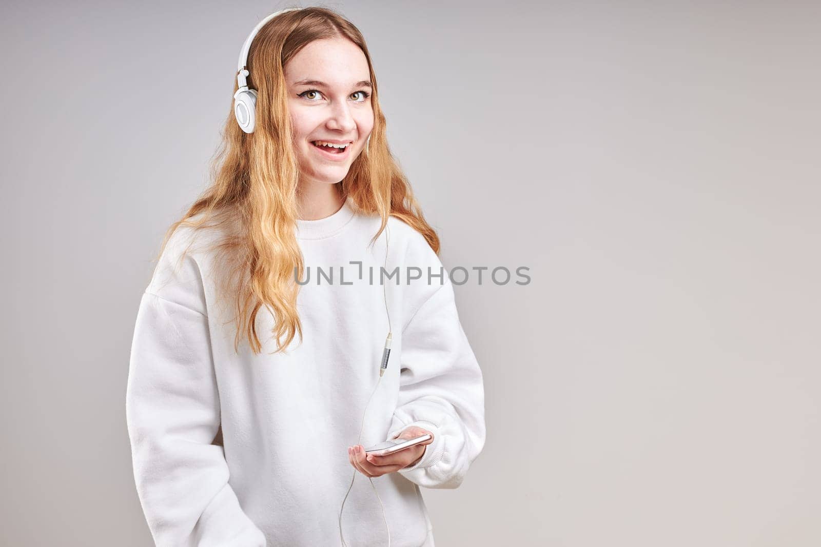 Young woman girl listening to music streaming content having fun watching video enjoying video chat talking with friends making gestures faces using smartphone earphones headphones standing over plain grey background