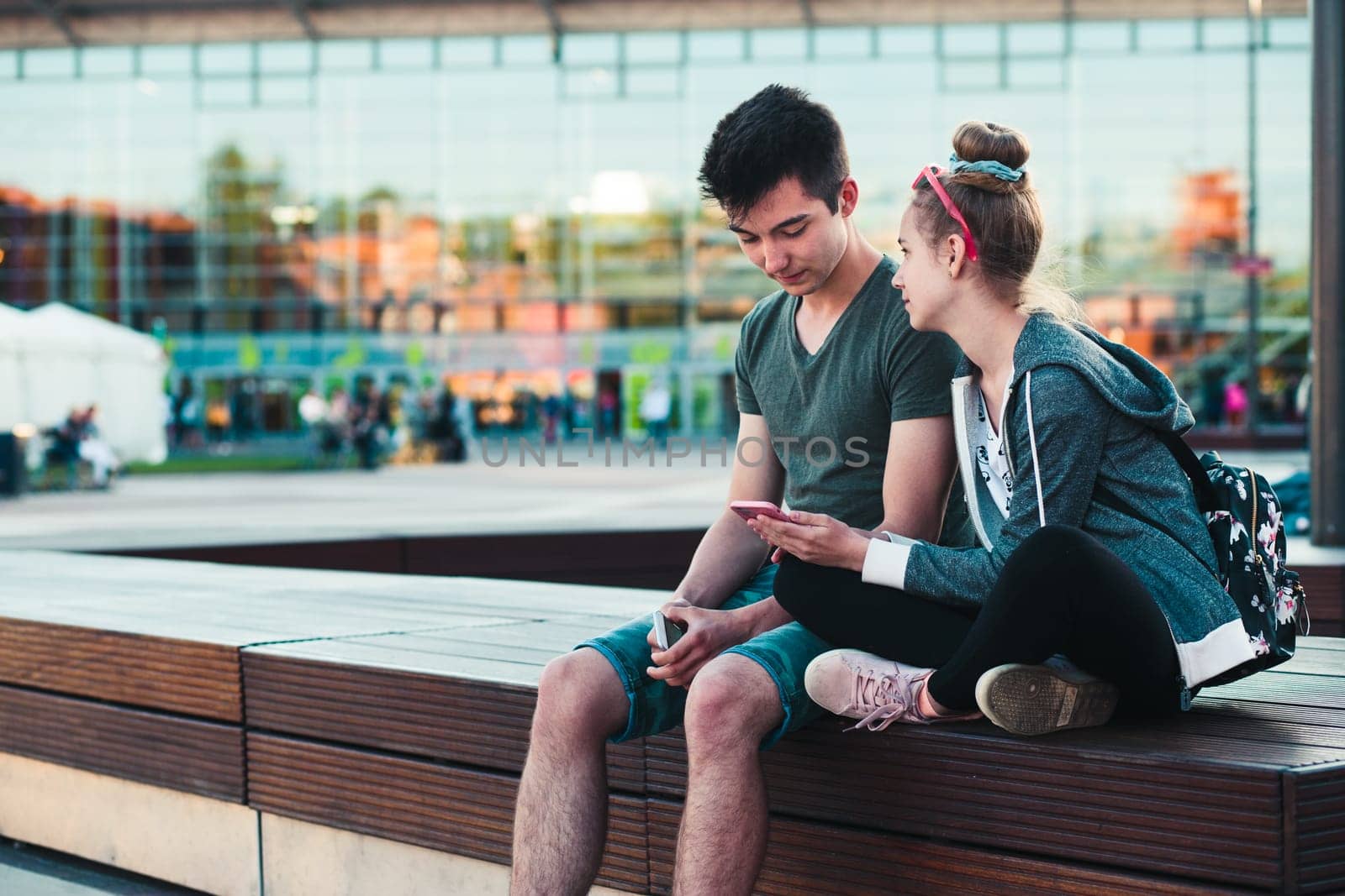 Couple of friends, teenage girl and boy,  having fun together with smartphones,  sitting in center of town, spending time together