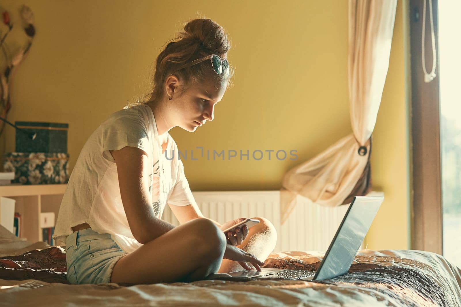 Young woman using portable computer and mobile phone, sitting on a bed, learning online at home. Candid people, real moments, authentic situations