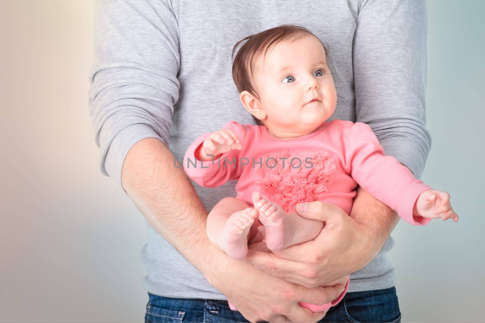 Baby sitting on fathers hands. Baby is wearing pink body suit and looking at right side