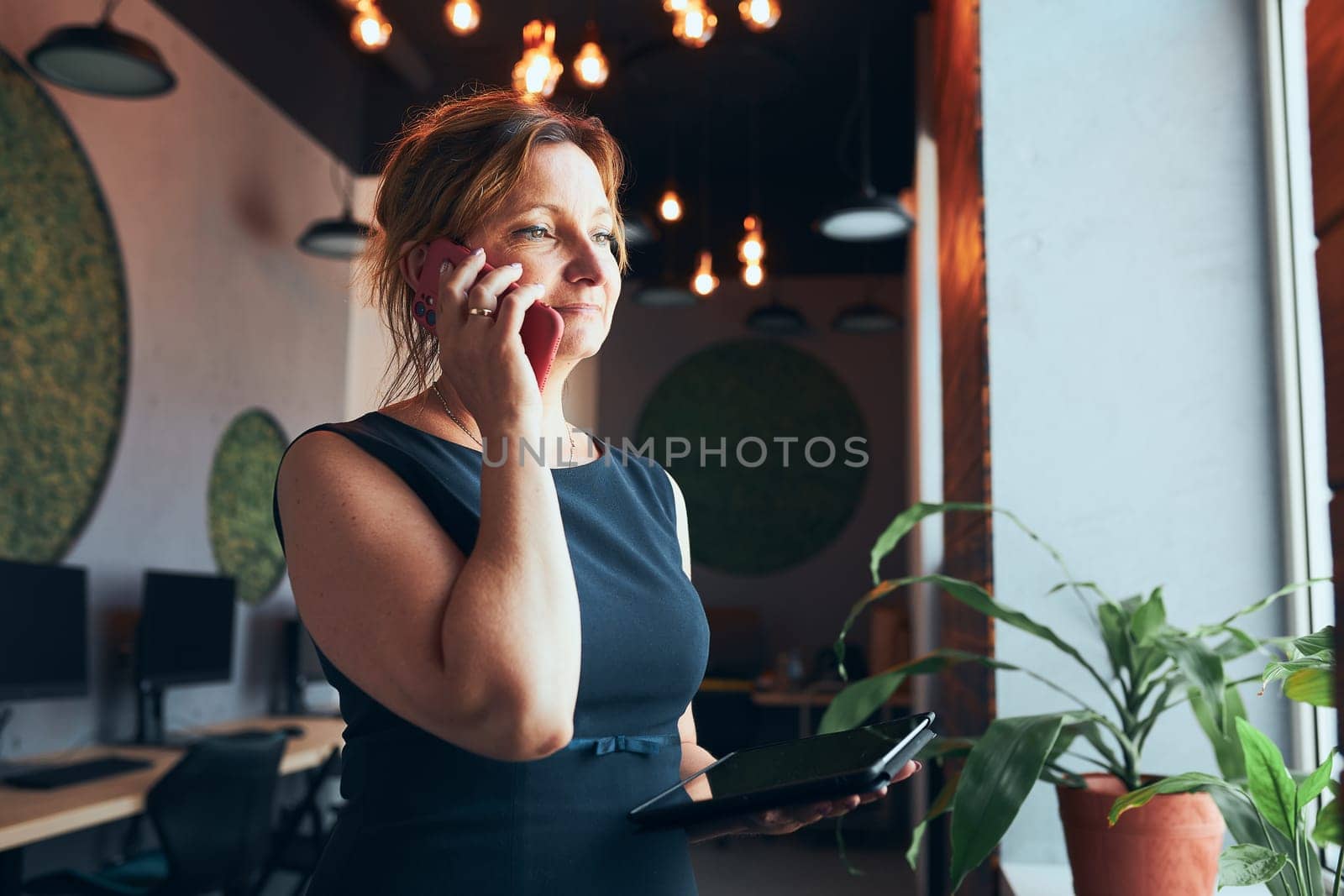 Businesswoman having business call working on tablet in office. Mature busy woman using touch pad computer standing by window in modern interior. Female manager focused on work holding digital device. Using technology