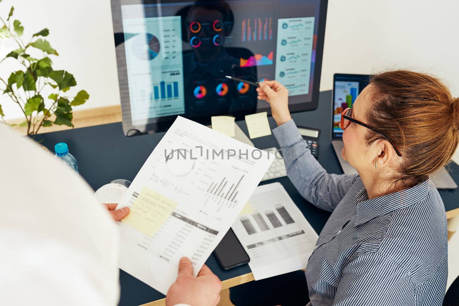 Woman entrepreneur discussing financial data with her colleague sitting at desk in office. Happy positive businesswoman working with charts and tables on computer. Two people working together. Workmates talking about project
