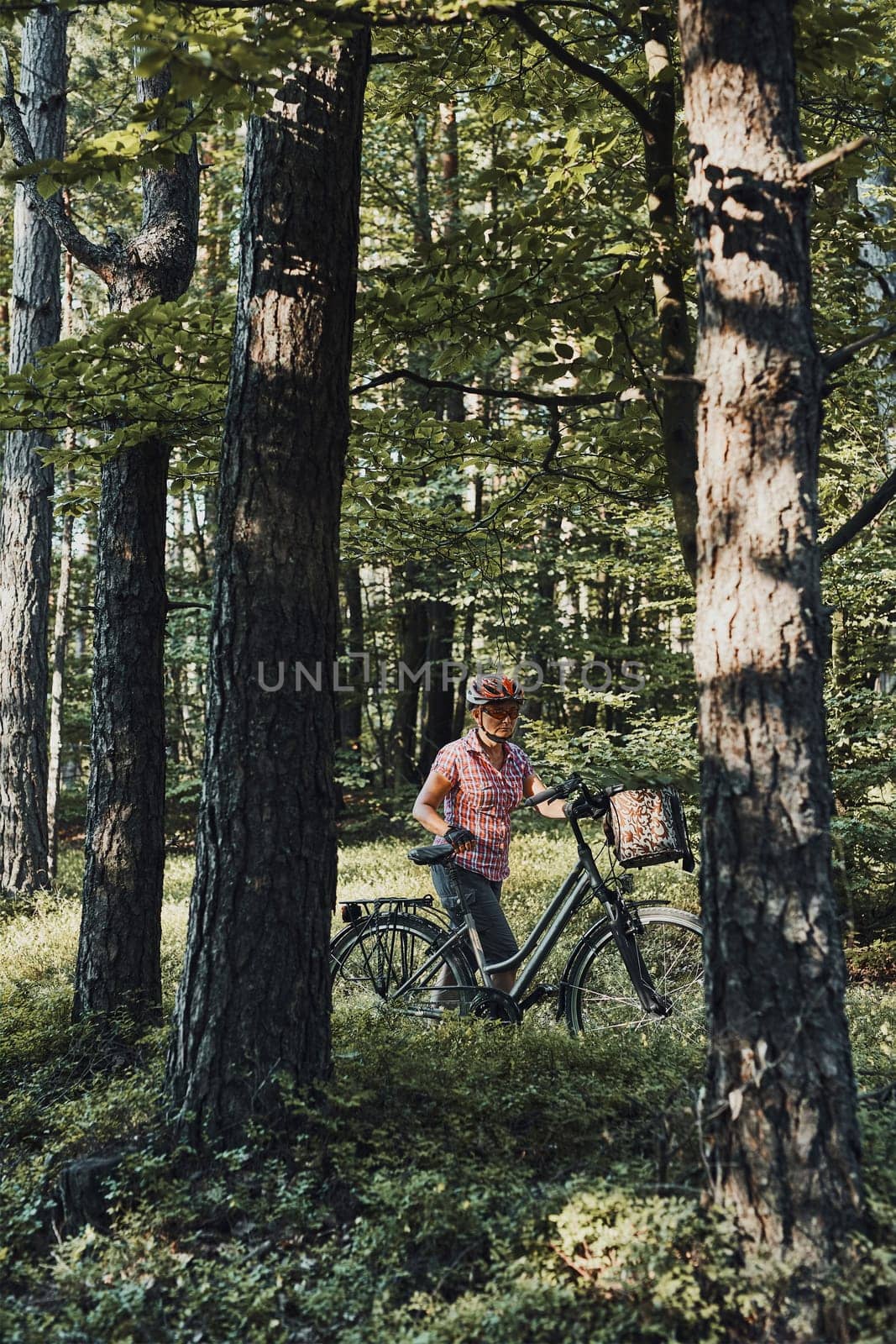 Active woman spending free summer vacation time on a bicycle trip in a forest. Woman wearing bicycle helmet and gloves holding bike with basket standing behind the trees