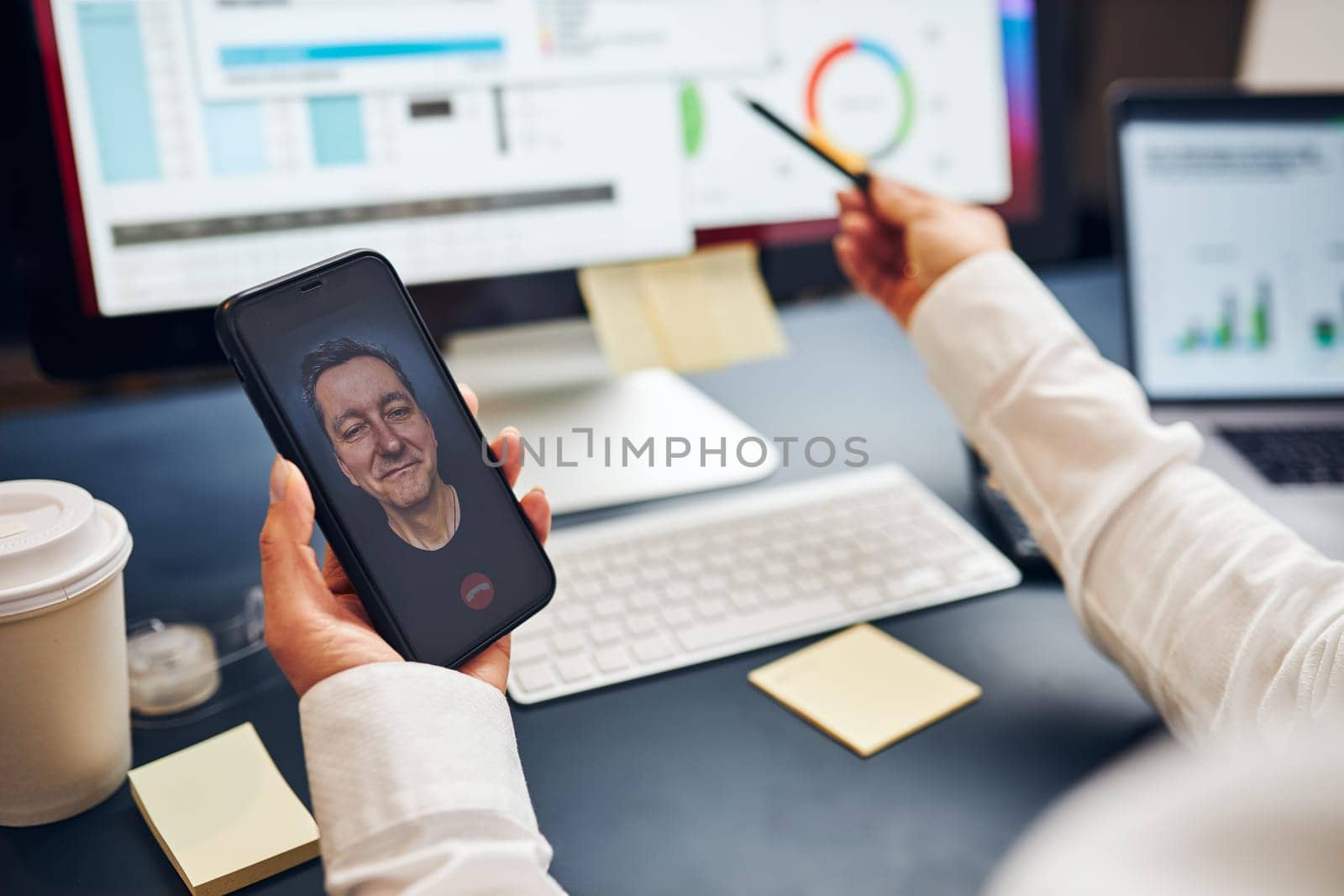 Woman entrepreneur having business video chat on smartphone with her business partner. Businesswoman working with data on charts, graphs and diagrams on computer screen. Woman holding smartphone sitting at desk in office