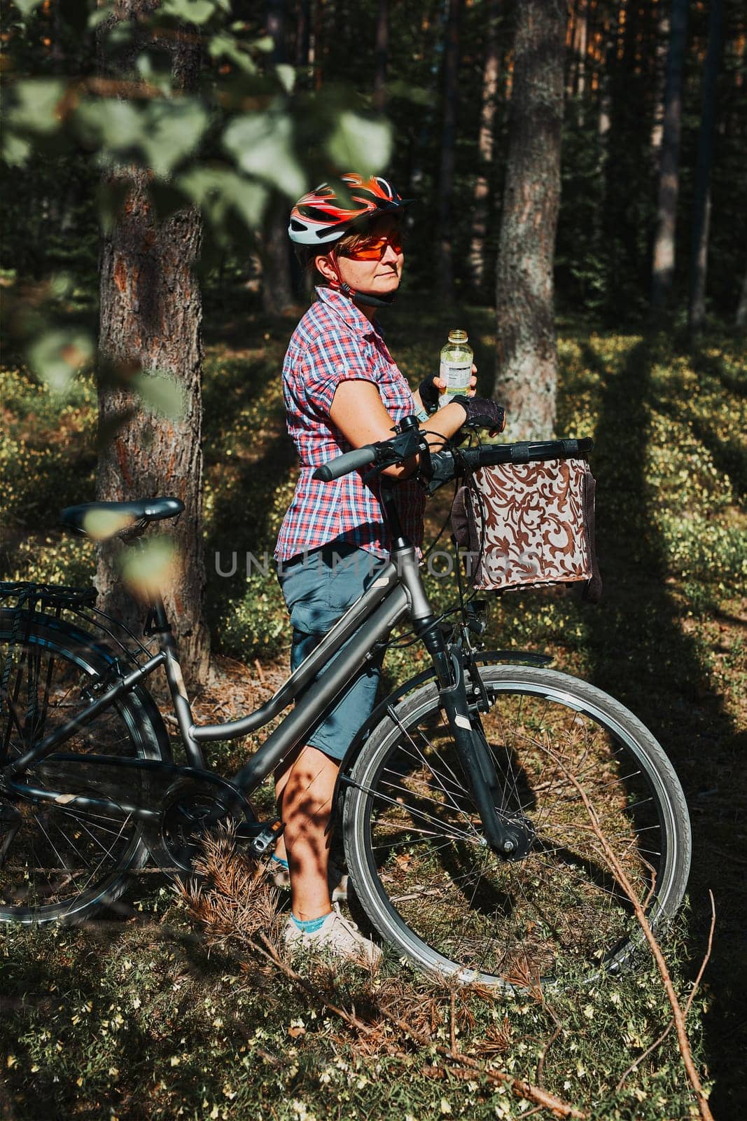 Active woman spending free summer vacation time on a bicycle trip in a forest. Woman wearing bicycle helmet and gloves holding bike with basket and drinking a water from bottle