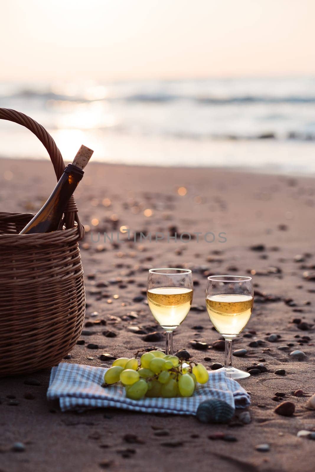 Two wine glasses with white wine standing on sand, on beach, beside grapes and wicker basket with bottle of wine. Sea waves in the background