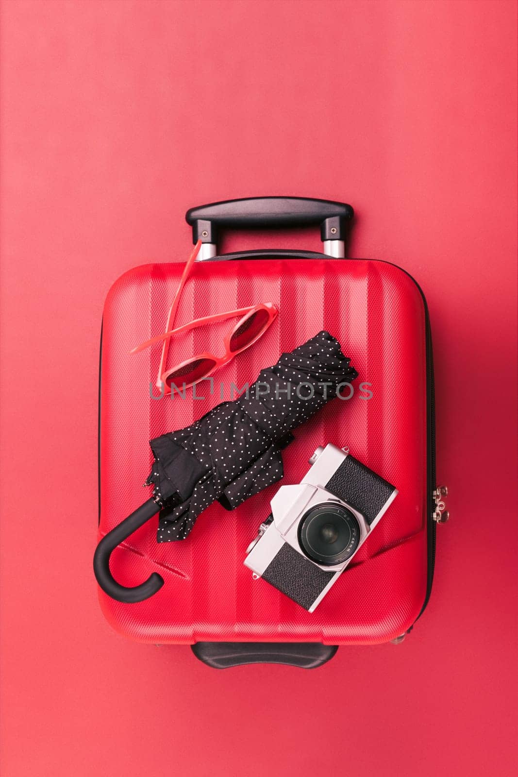 Red travel suitcase with red sunglasses, old camera, black umbrella on red background. Minimal style