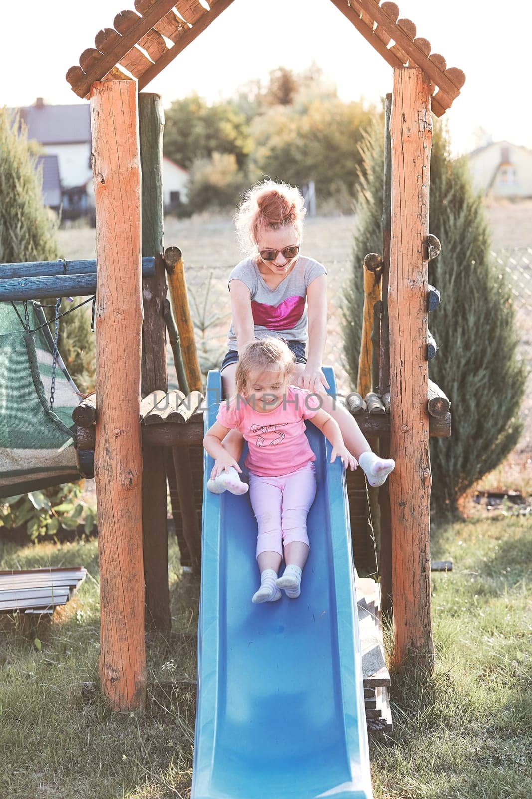 Teenage girl playing with her younger sister in a home playground in a backyard. Happy smiling sisters having fun on a slide together on summer day. Real people, authentic situations