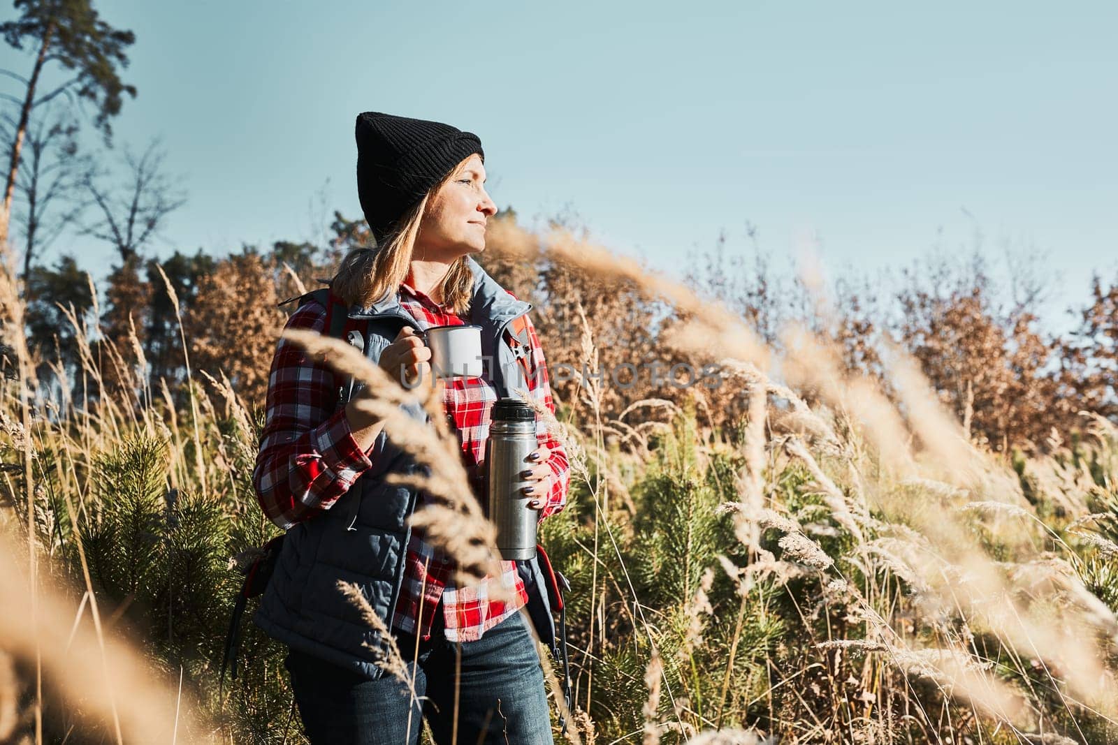 Woman taking break and relaxing with cup of coffee during summer trip. Woman standing on trail and looking away. Female with backpack hiking through tall grass along path in mountains. Spending summer vacation close to nature