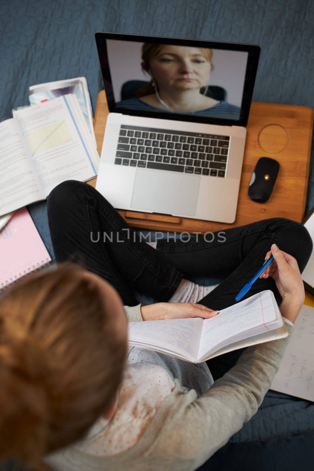 Young woman student having classes, learning online, watching lesson remotely, listening to professor, talking with classmates on video call from home during quarantine. Young girl using laptop, headphones, books, manuals sitting on bed
