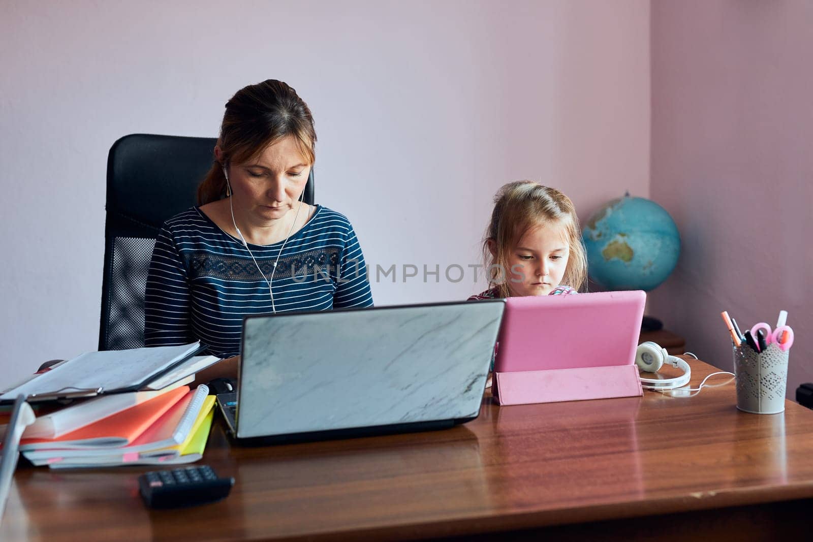 Woman mother working doing her job remotely during video chat call stream online course webinar on laptop from home while her daughter watching video on tablet. Woman sitting at desk in front of computer looking at screen