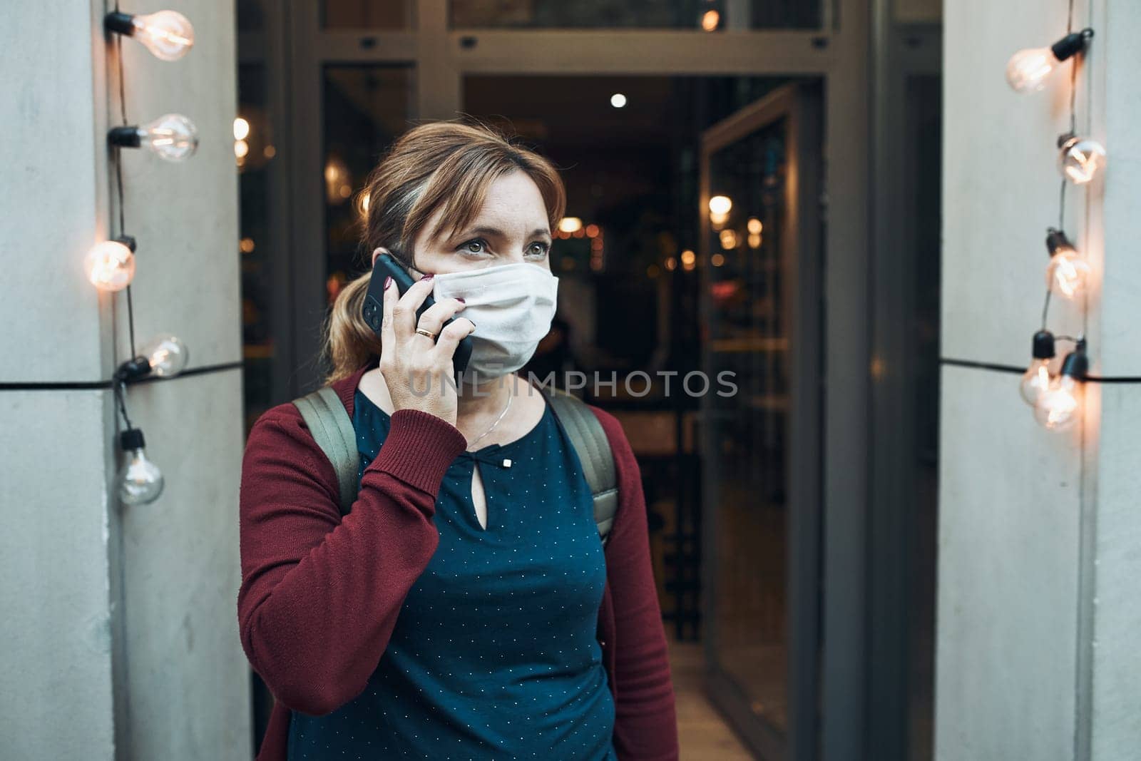 Young woman talking on phone standing in a front of restaurant downtown wearing the face mask to avoid virus infection and to prevent the spread of disease in time of coronavirus