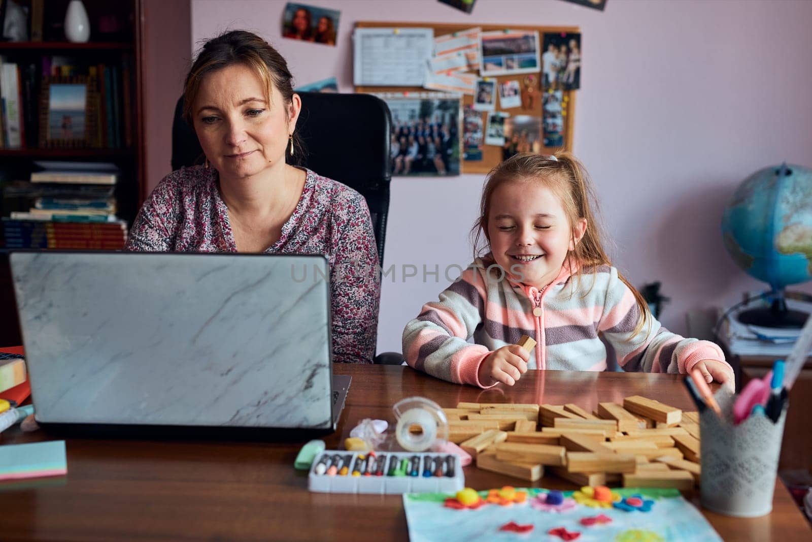 Woman mother working doing her job remotely during video chat call stream online course webinar on laptop from home while her daughter playing with bricks toy. Woman sitting at desk in front of computer looking at screen