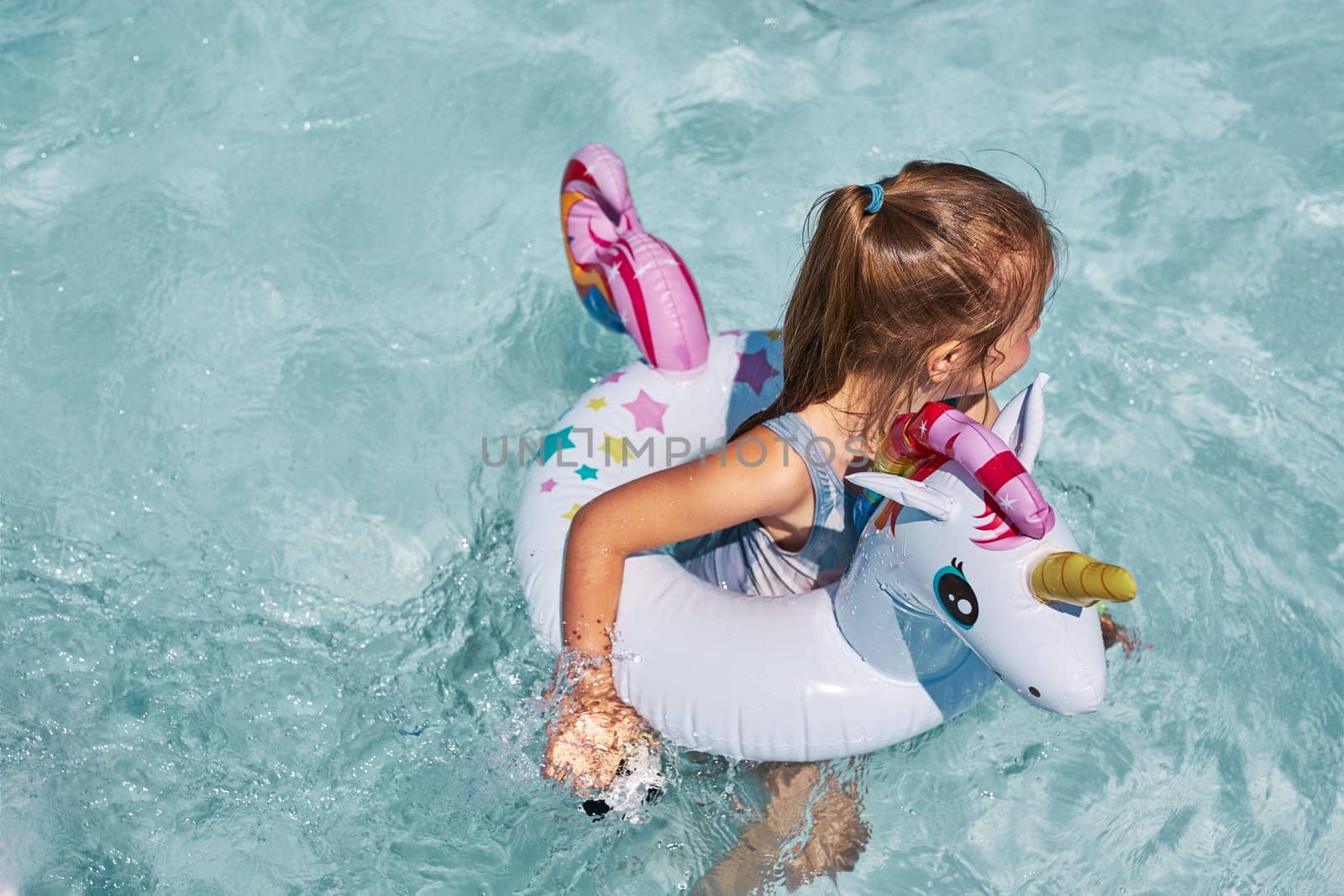 Little girl playing in a swimming pool with inflatable rubber ring toy in the shape of unicorn. View from above of kid in a turquoise water swimming in rubber colorful ring