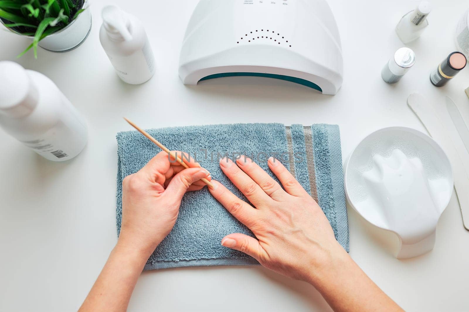 Woman preparing nails to apply gel hybrid polish using UV lamp. Beauty wellness spa treatment concept. Cosmetic products, UV lamp, green leaves on white table. Spa, manicure, skin care concept. Flat lay, overhead view