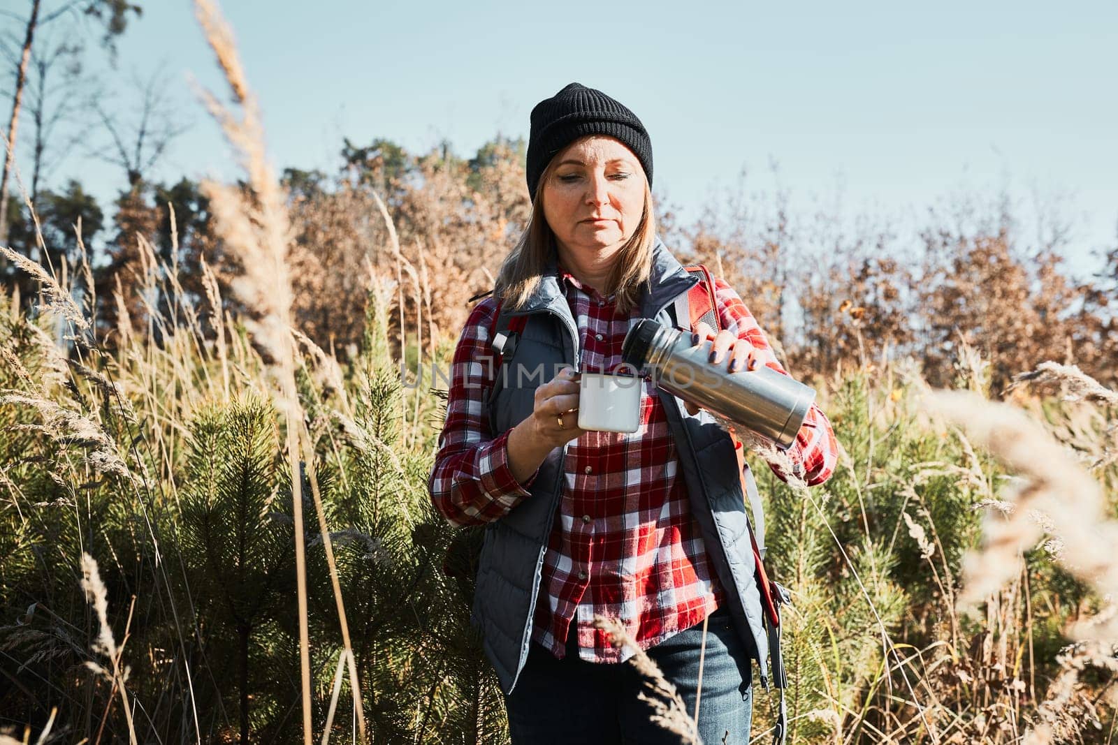 Woman taking break and relaxing with cup of coffee during summer trip. Woman standing on trail and looking away. Female with backpack hiking through tall grass along path in mountains. Spending summer vacation close to nature