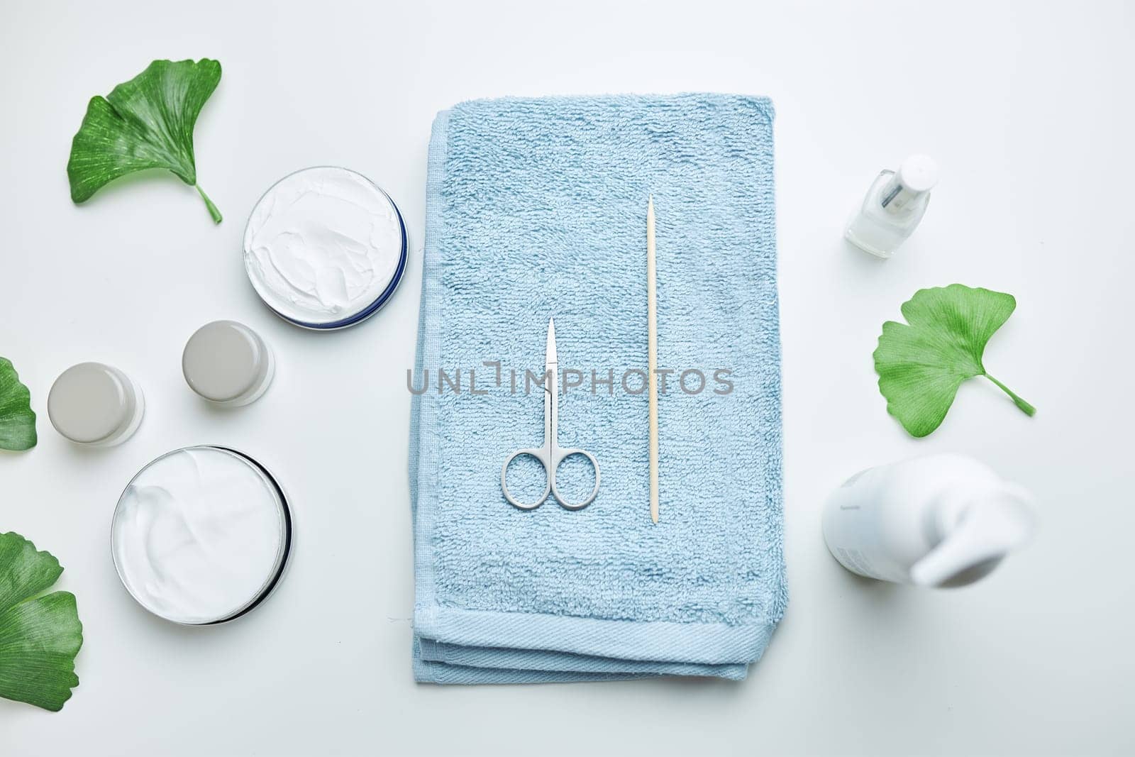 Cosmetic products set, green leaves, blue towel on white table. Spa, manicure, skin care concept. Flat lay, overhead view