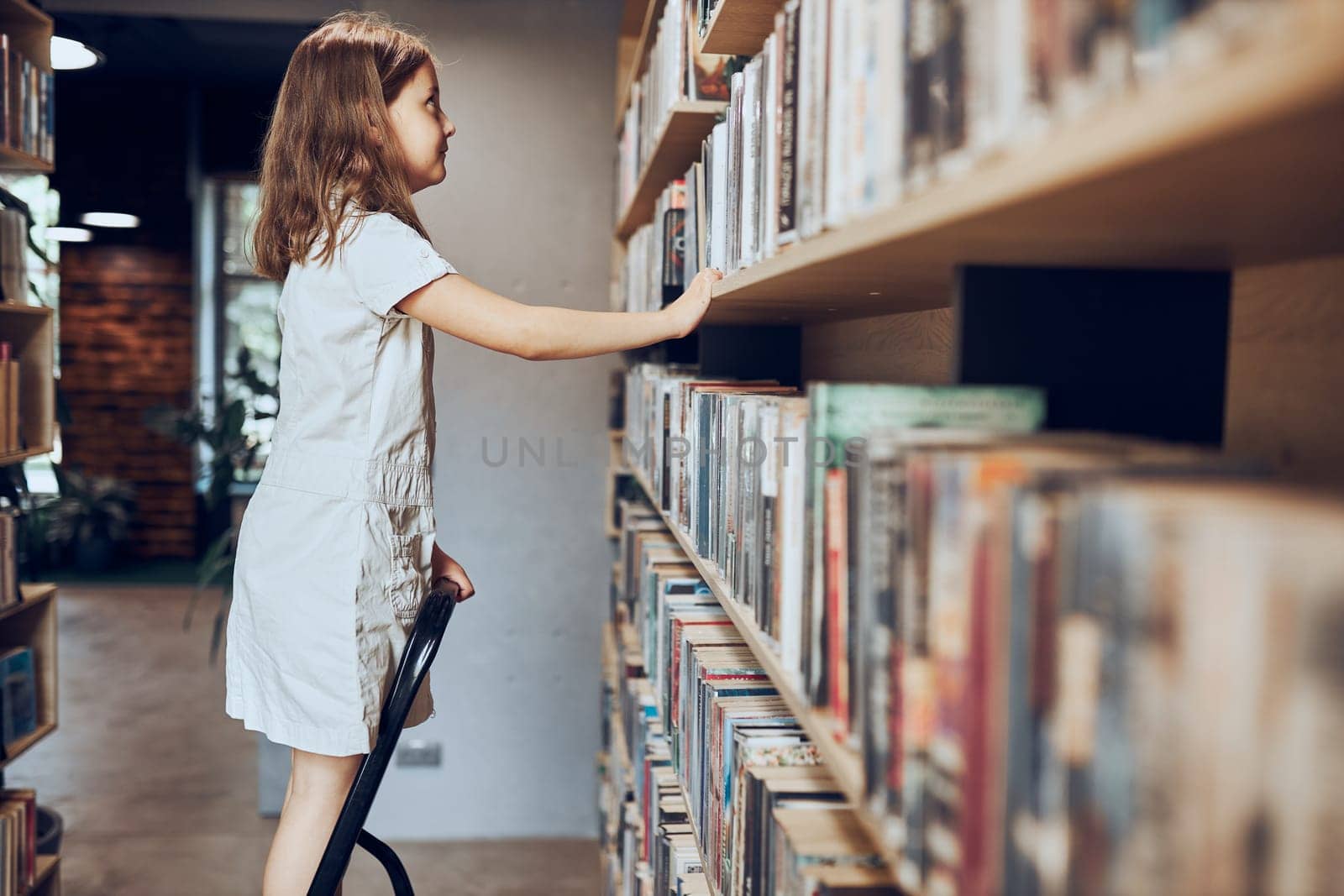 Schoolgirl looking at books on top bookshelf in school library. Smart girl selecting literature for reading. Books on shelves in bookstore. Learning from books. School education. Benefits of everyday reading. Child curiosity. Back to school