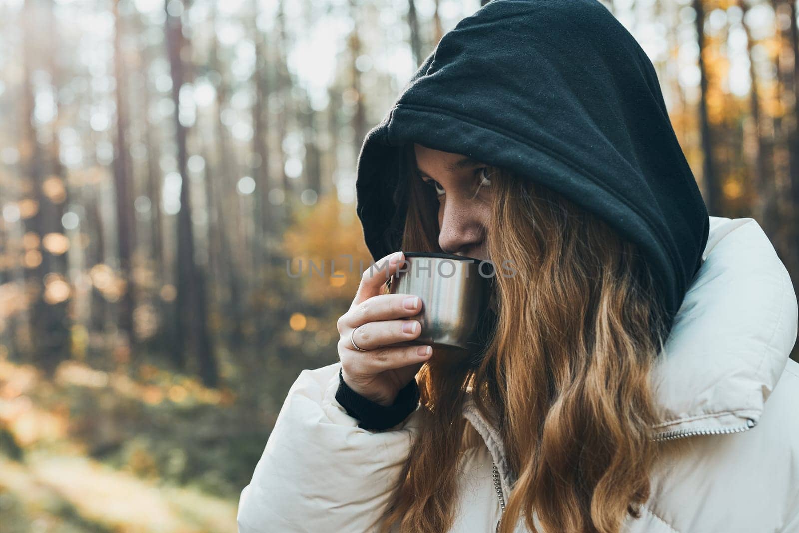 Woman in a hood having break during autumn trip holding cup with hot drink from thermos flask on autumn cold day. Active girl wandering in a forest actively spending time
