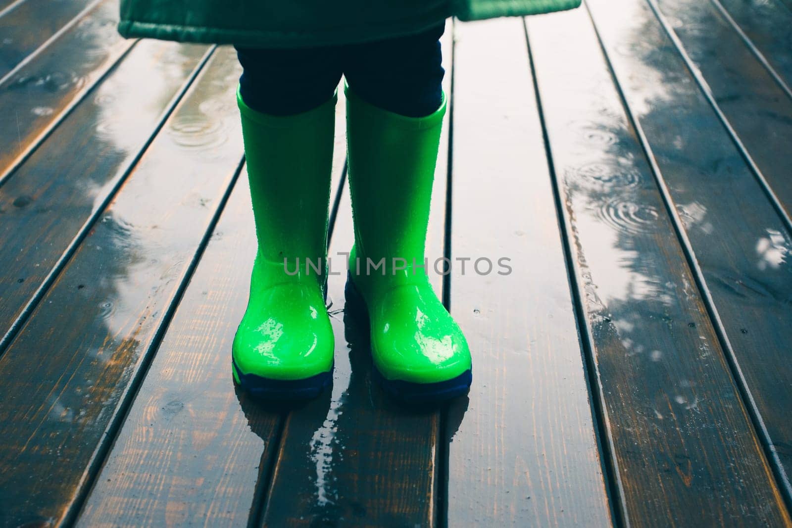 Closeup of legs of kid standing on a porch wearing green wellies and raincoat. Rain boots in bright green color