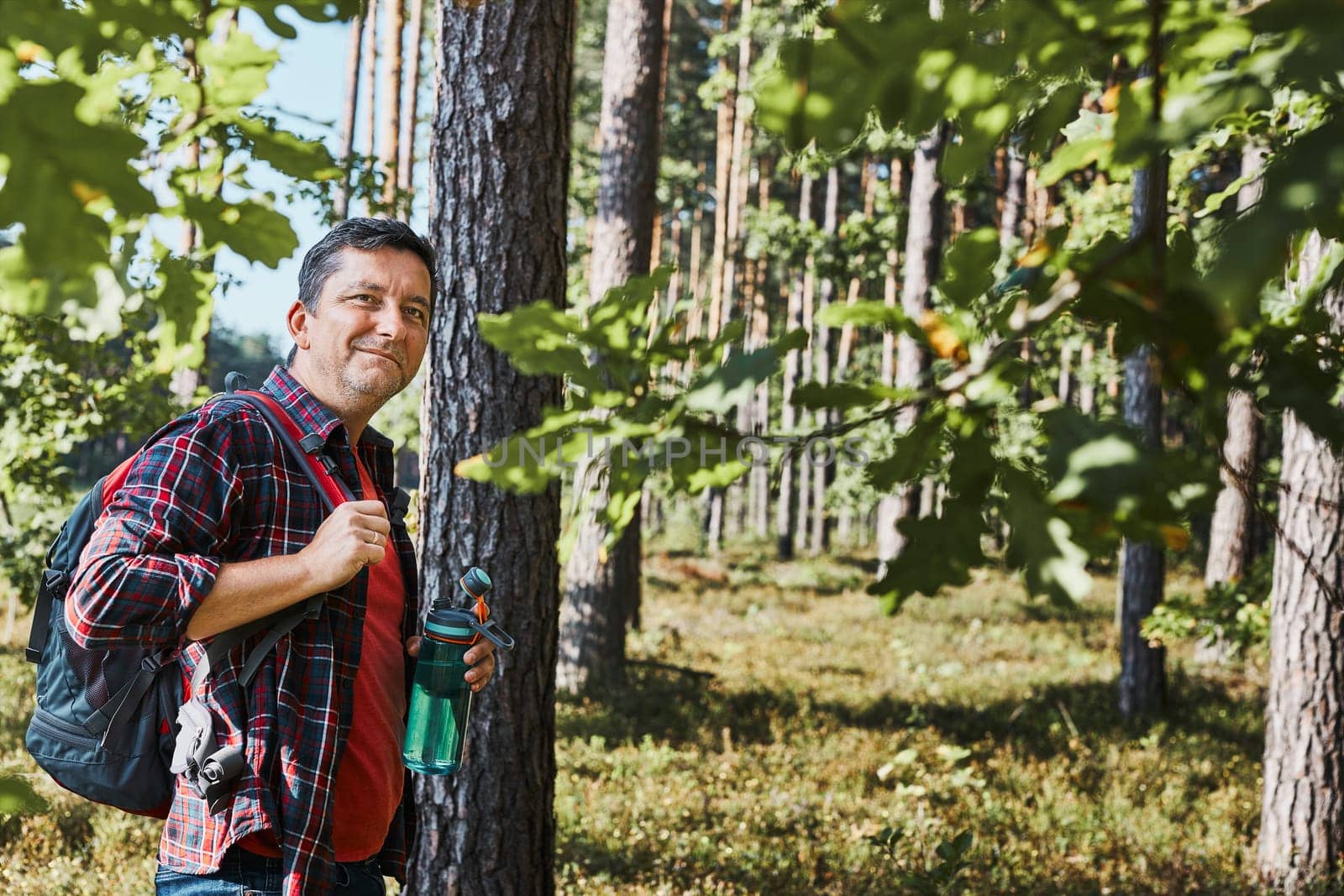 Man with backpack hiking in forest, actively spending summer vacation close to nature. Man walking on path among trees, holding bottle of water, enjoying hike on sunny vacation day