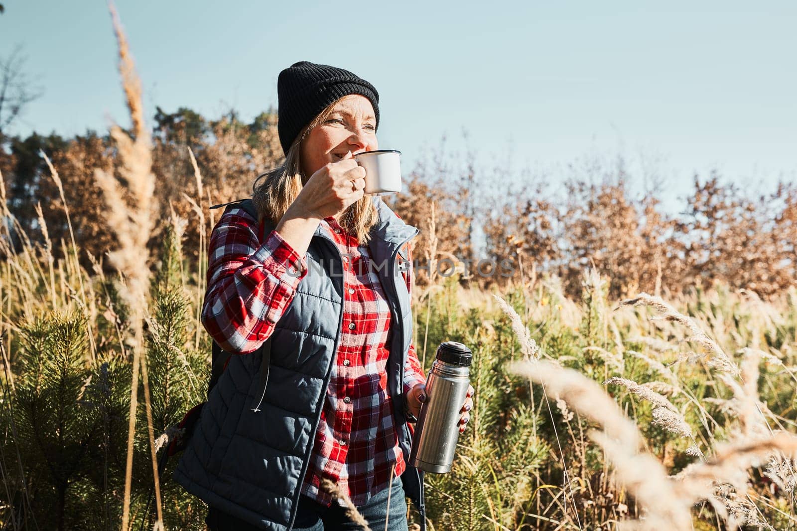 Woman taking break and relaxing with cup of coffee during summer trip. Woman standing on trail and looking away. Woman with backpack hiking through tall grass along path in mountains. Spending summer vacation close to nature