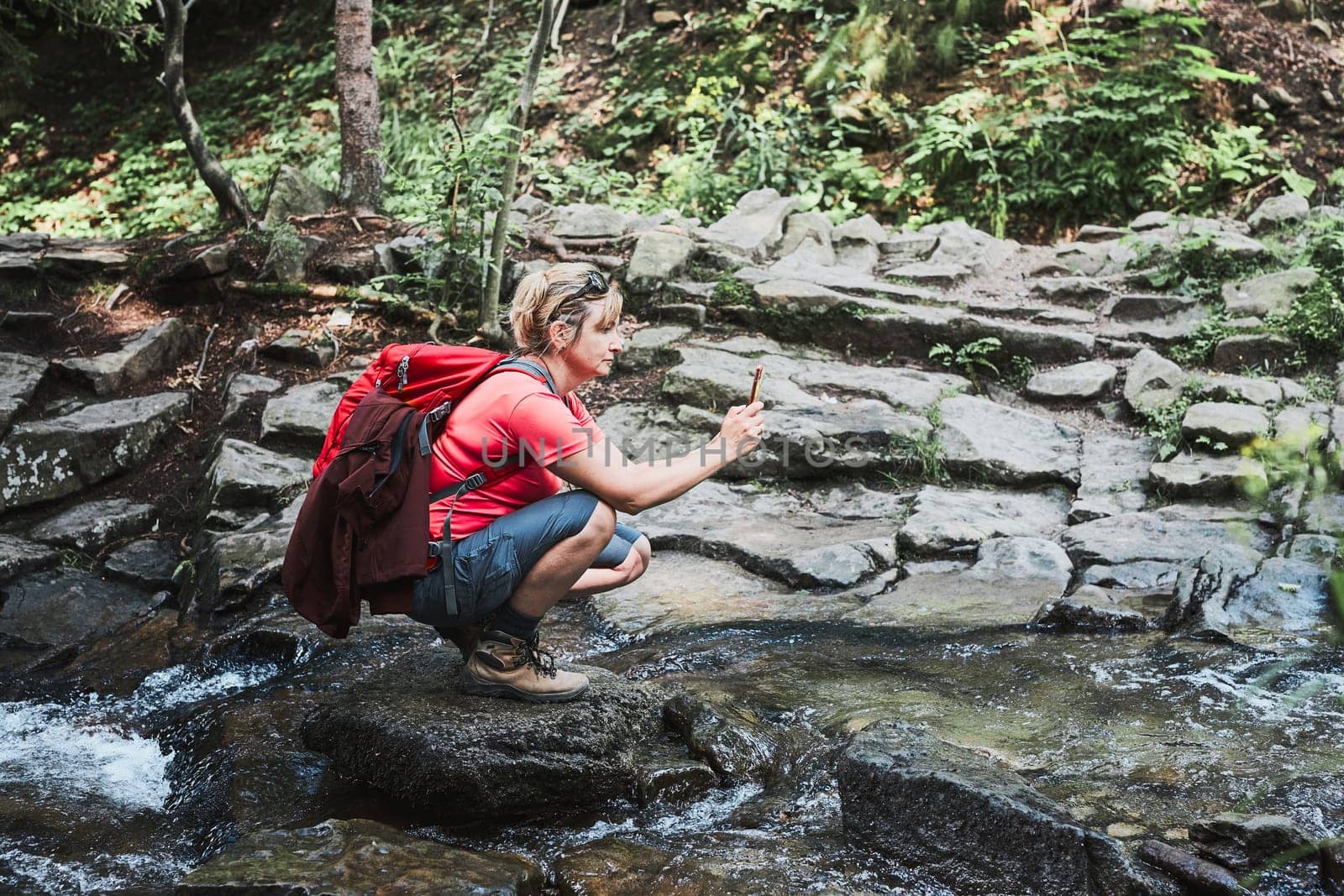 Taking pictures from vacation. Woman with backpack taking photos of landscape using smartphone camera standing on rock on mountain stream during summer trip