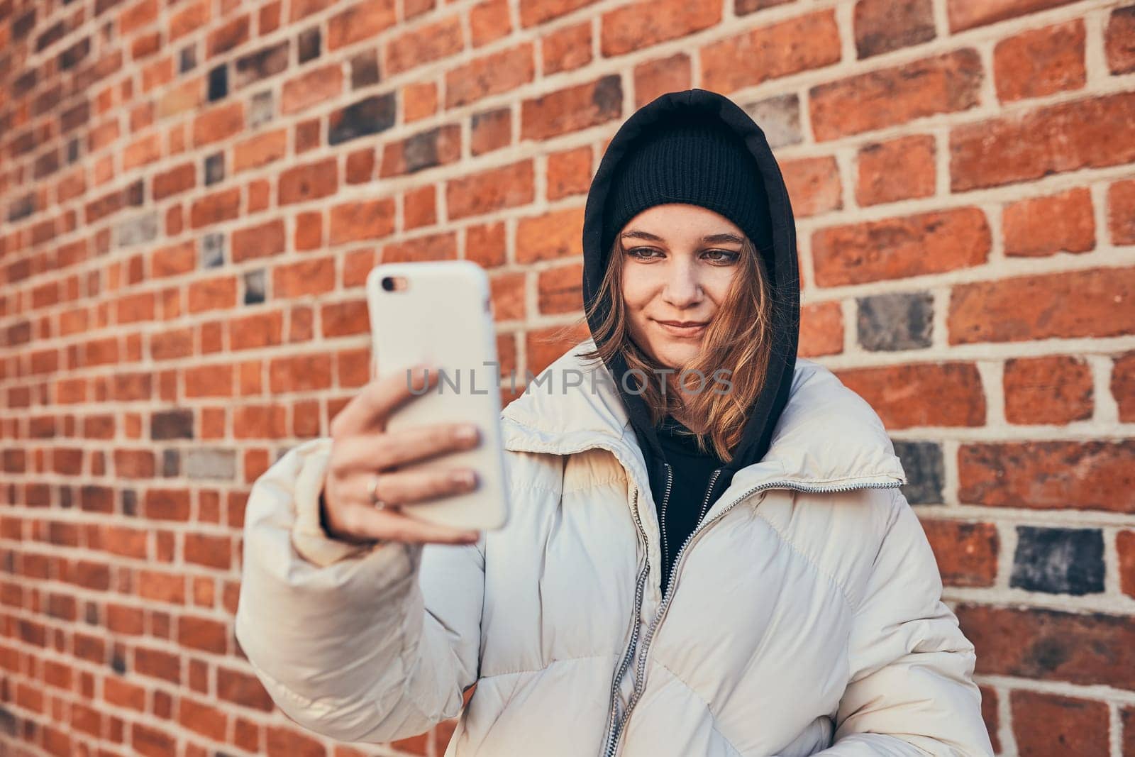 Young woman having video call, talking remotely, taking selfie photo holding smartphone standing at brick wall