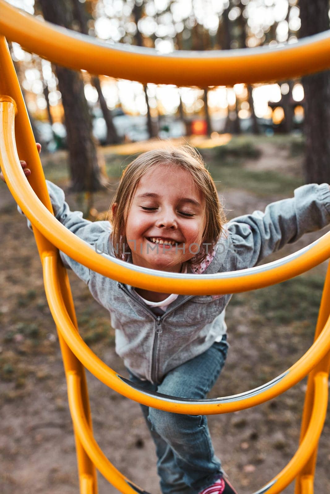 Playful happy girl preschooler playing on a playground climbing on monkey bars smiling with closed eyes. Child having fun playing outdoors in a park on summer day