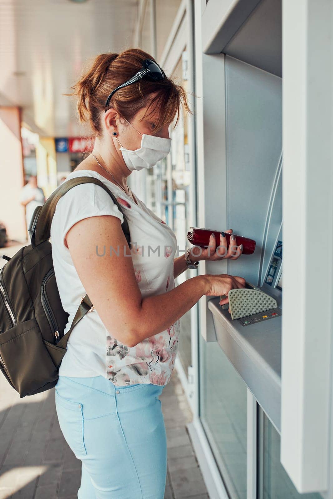 Woman withdrawing money from atm machine using debit or credit card standing outdoors in the street, wearing face mask to cover mouth and nose during pandemic coronavirus outbreak