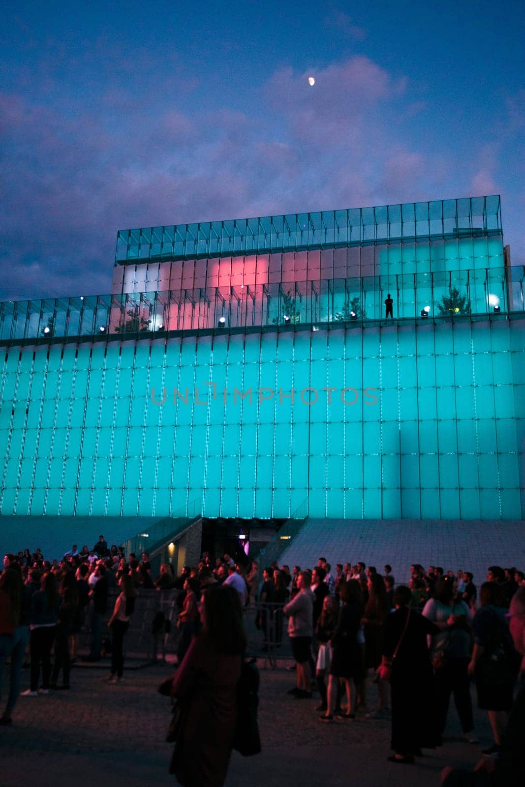 Festival event party concert with people standing on a square in a city center at night. Backlit glass construction above them