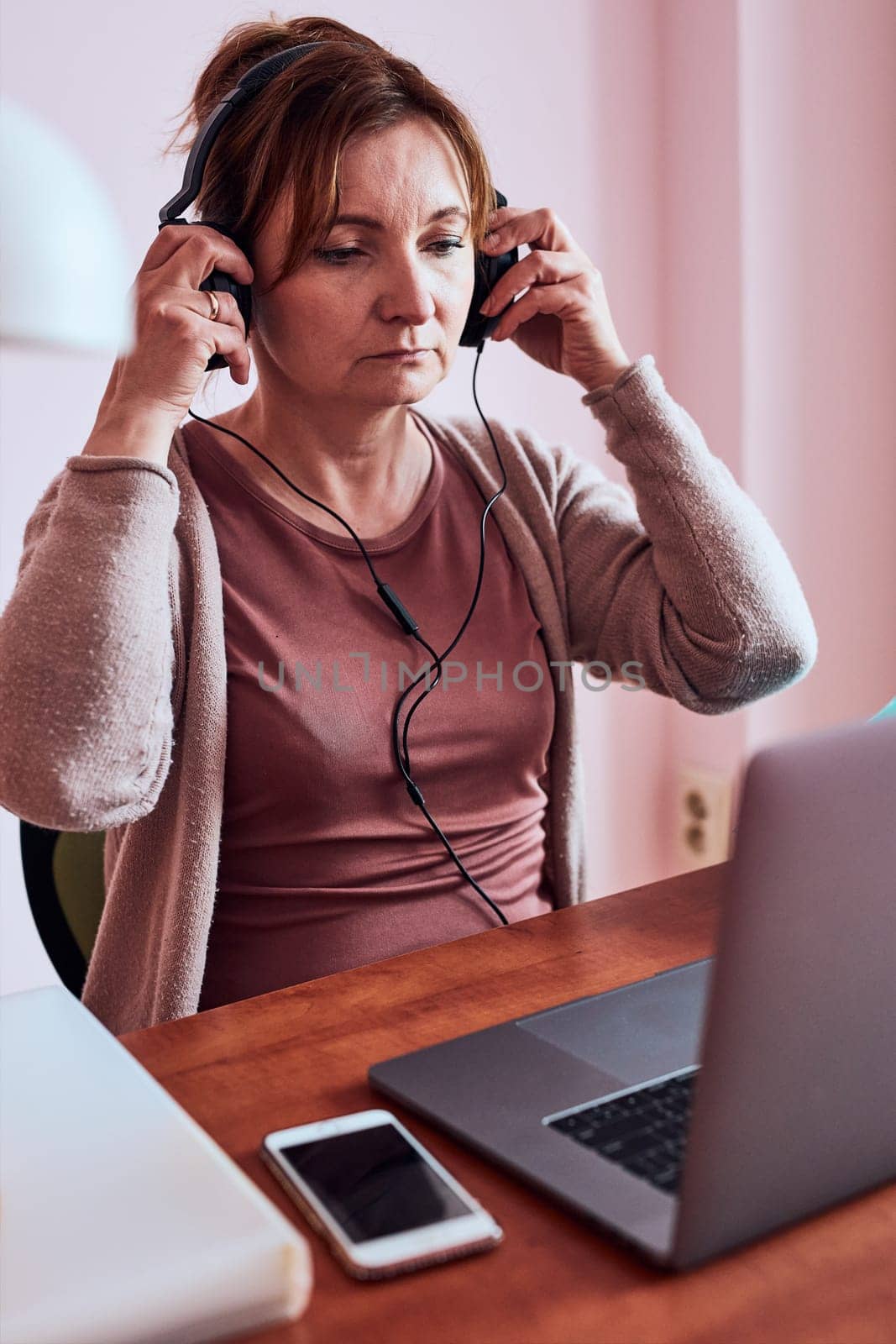 Woman working talking doing her job remotely during video chat phone call on laptop from home. Woman sitting at desk in front of computer looking at screen using headphones and smartphone. Concept of remote working