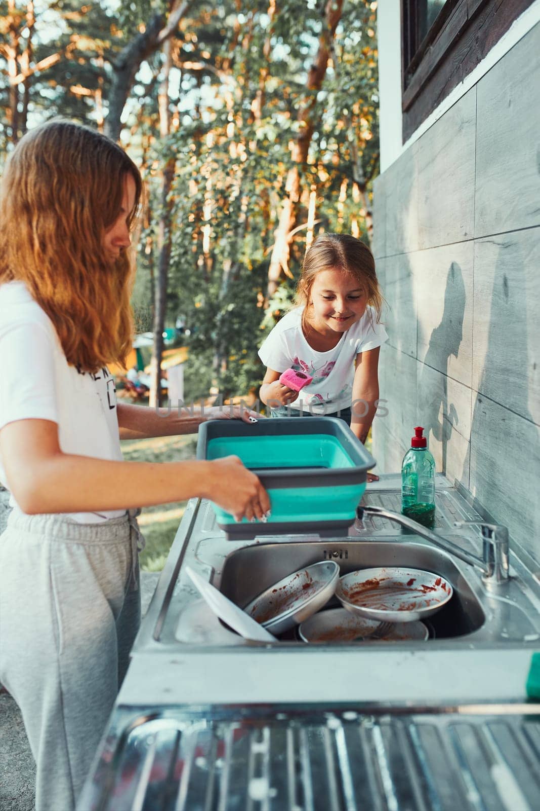 Teenager girl washing up the dishes pots and plates with help her younger sister in the outdoor kitchen during vacations on camping. Camp life. Sisters spending time and working together