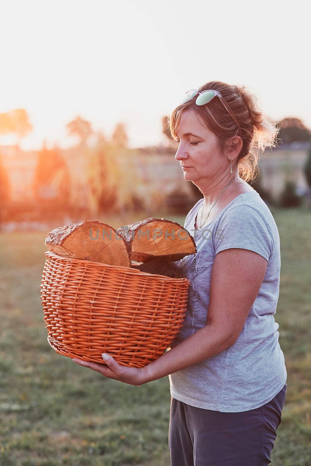 Adult woman putting the wood logs to wicker basket, standing by wood shed in the backyard. Real people, authentic situations