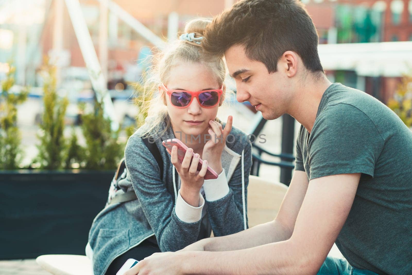 Couple of friends, teenage girl and boy,  having fun using smartphones sitting in center of town, spending time together