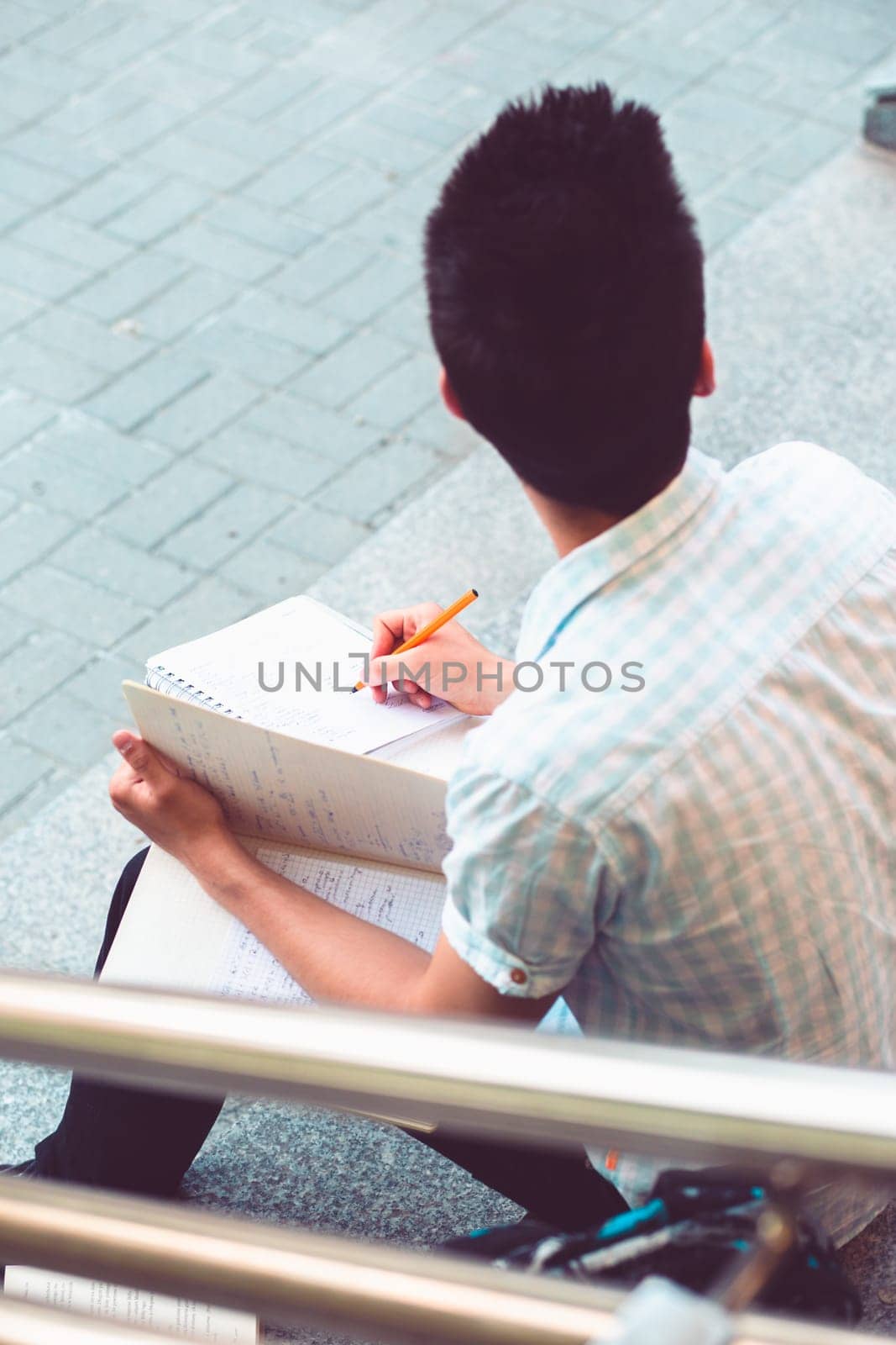 Student making a notes sitting on stairs outside of university. Young boy wearing blue shirt and dark jeans.
