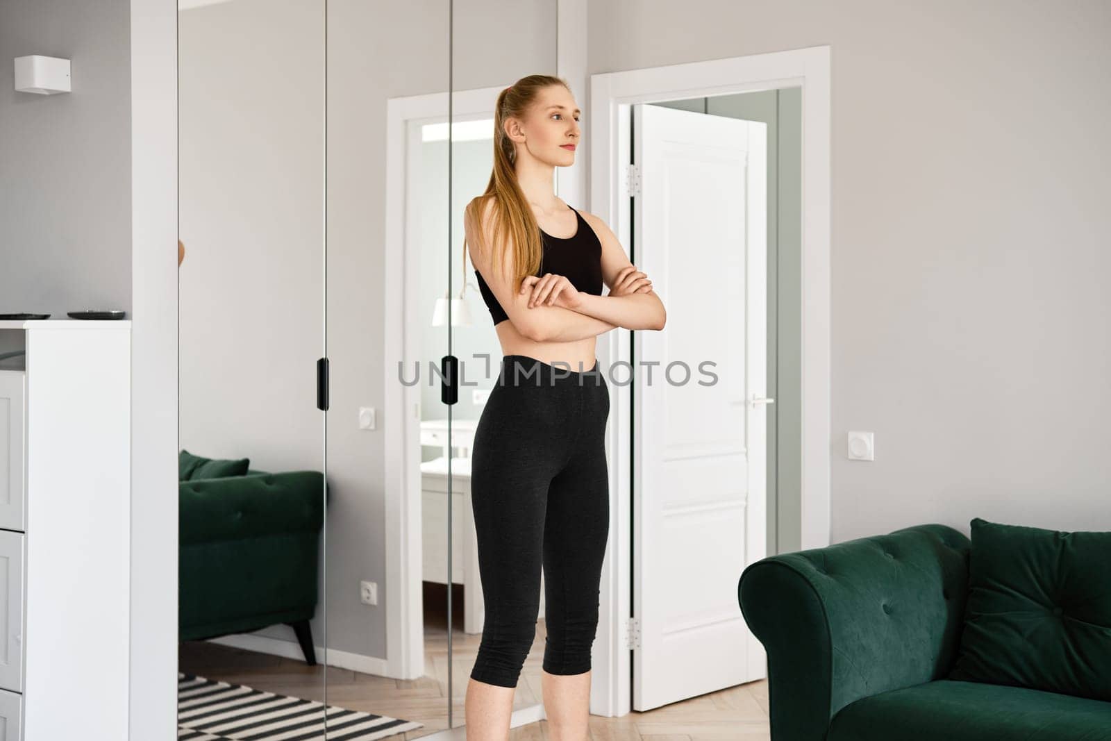 Young woman in sportswear stands in living room with her arms crossed on her chest