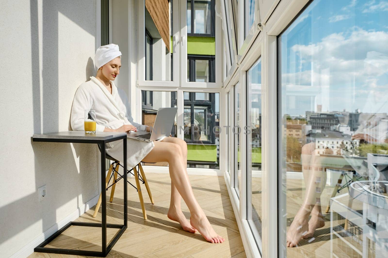 Barefoot woman in a bathrobe sits on balcony of her new high-rise apartment and using laptop