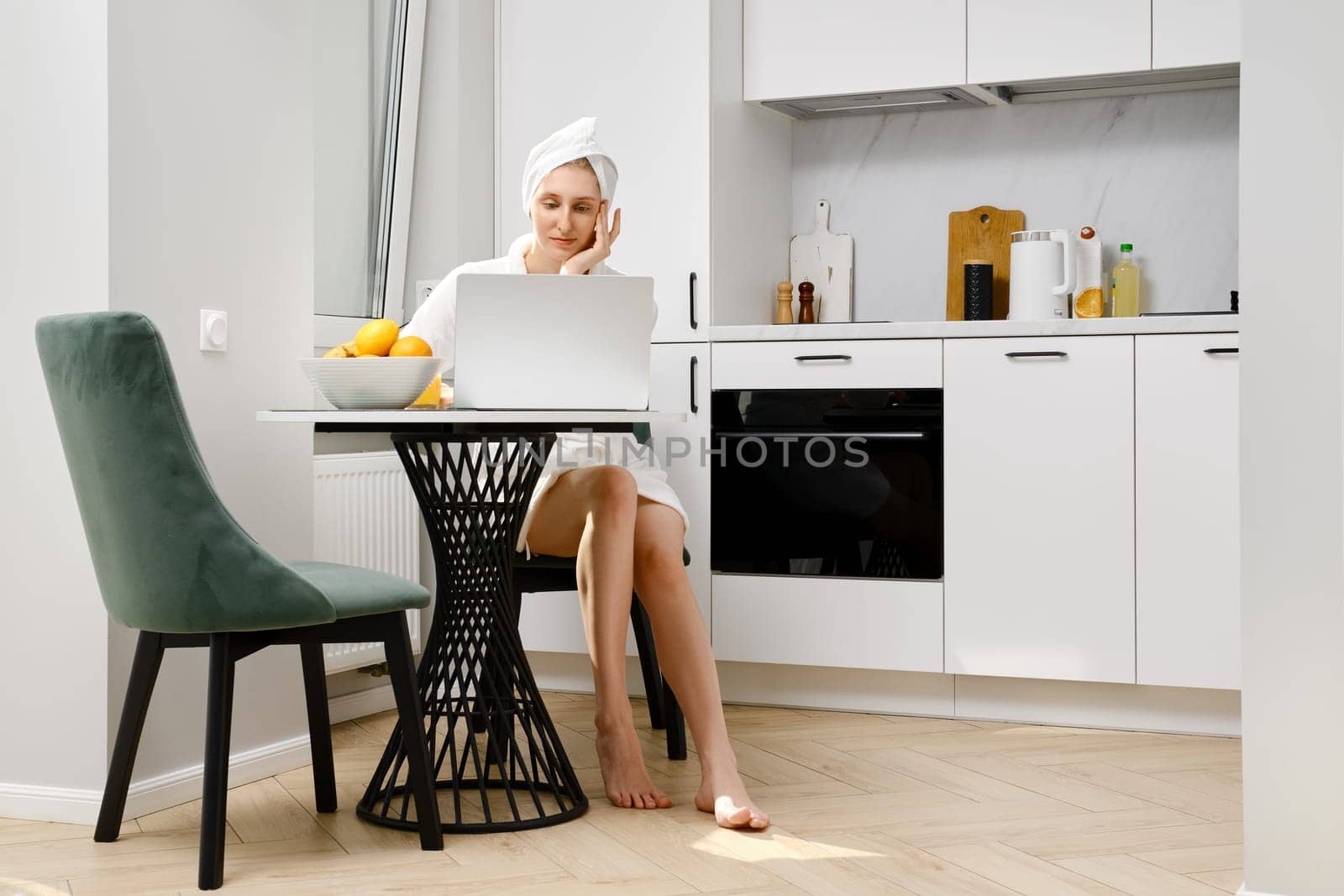 Young barefoot woman in bathrobe sits in the kitchen and browsing social media on laptop