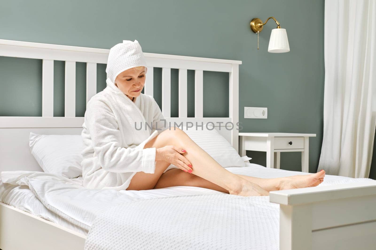 Senior woman sitting on bed and applying lotion on her feet after shower